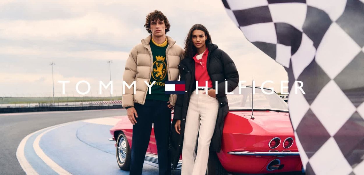 Dos modelos están de pie en una pista de carreras. El hombre lleva una chaqueta acolchada beige sobre un suéter verde. La mujer viste un abrigo acolchado negro y una parte superior roja.