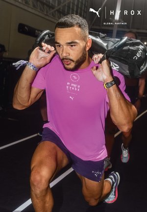 Uomo con camicia rosa esegue affondi con pesi al chiuso, portando una borsa pesante sulle spalle, con espressione concentrata durante un evento di fitness.
