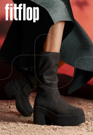 Schwarze Plateau-Stiefel bis zur Mitte der Wade, getragen mit einem gerippten Rock vor rotem Hintergrund, stehend auf strukturiertem Boden mit dem Schriftzug "fitflop".