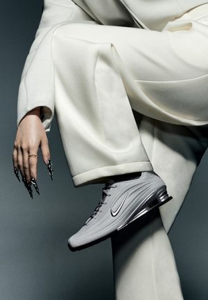 Zapatillas Nike grises con tejido texturizado, acentos plateados y un tacón elevado. Se llevan con pantalones anchos blancos en una pose minimalista.