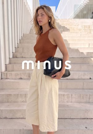 Jonge vrouw in een bruin mouwloos topje en crème broek met wijde pijpen, houdend een zwarte clutch, staand op buitenstenen trappen met een moderne witte reling.