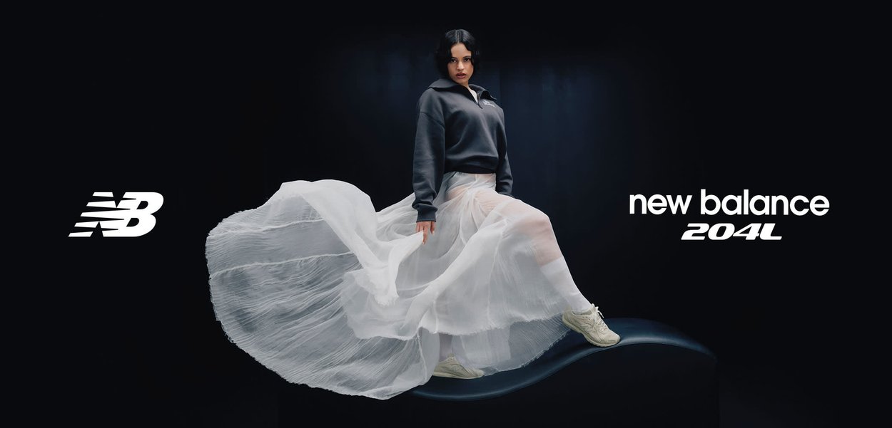 Mujer con falda blanca fluida y sudadera oscura, calzando zapatillas beige New Balance 204L, posando sobre una superficie negra curva contra un fondo oscuro.