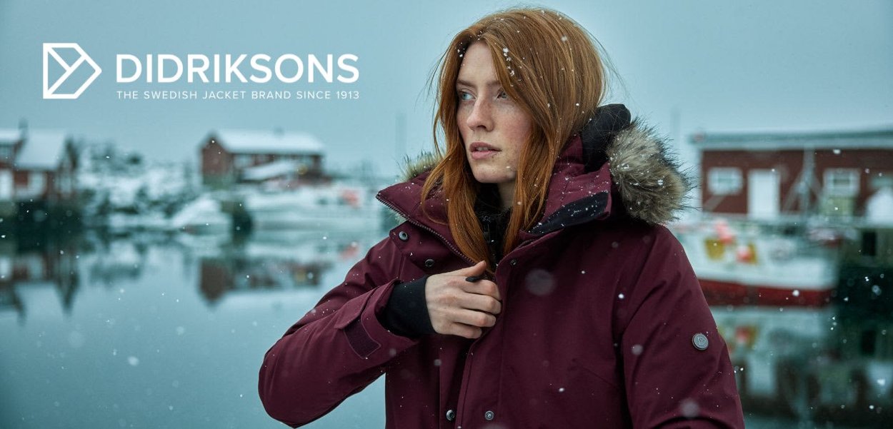 Frau in einer kastanienbraunen Winterjacke mit fellbesetzter Kapuze, die neben einem verschneiten Ufer steht, mit unscharfen Gebäuden im Hintergrund.