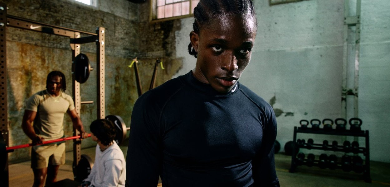 Haut de sport noir à manches longues avec une coupe ajustée et une texture lisse. En arrière-plan, on aperçoit du matériel de gym, notamment une barre d'haltères et des poids.