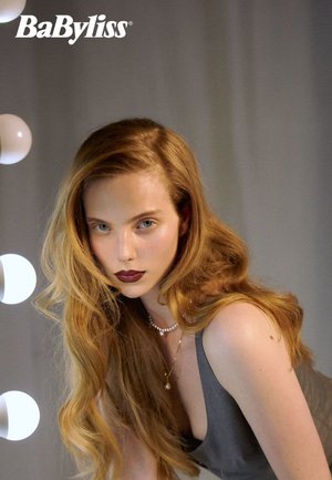 Model with long, wavy blonde hair in a grey top. She wears a pearl necklace and dark lipstick, with soft lighting from round bulbs.