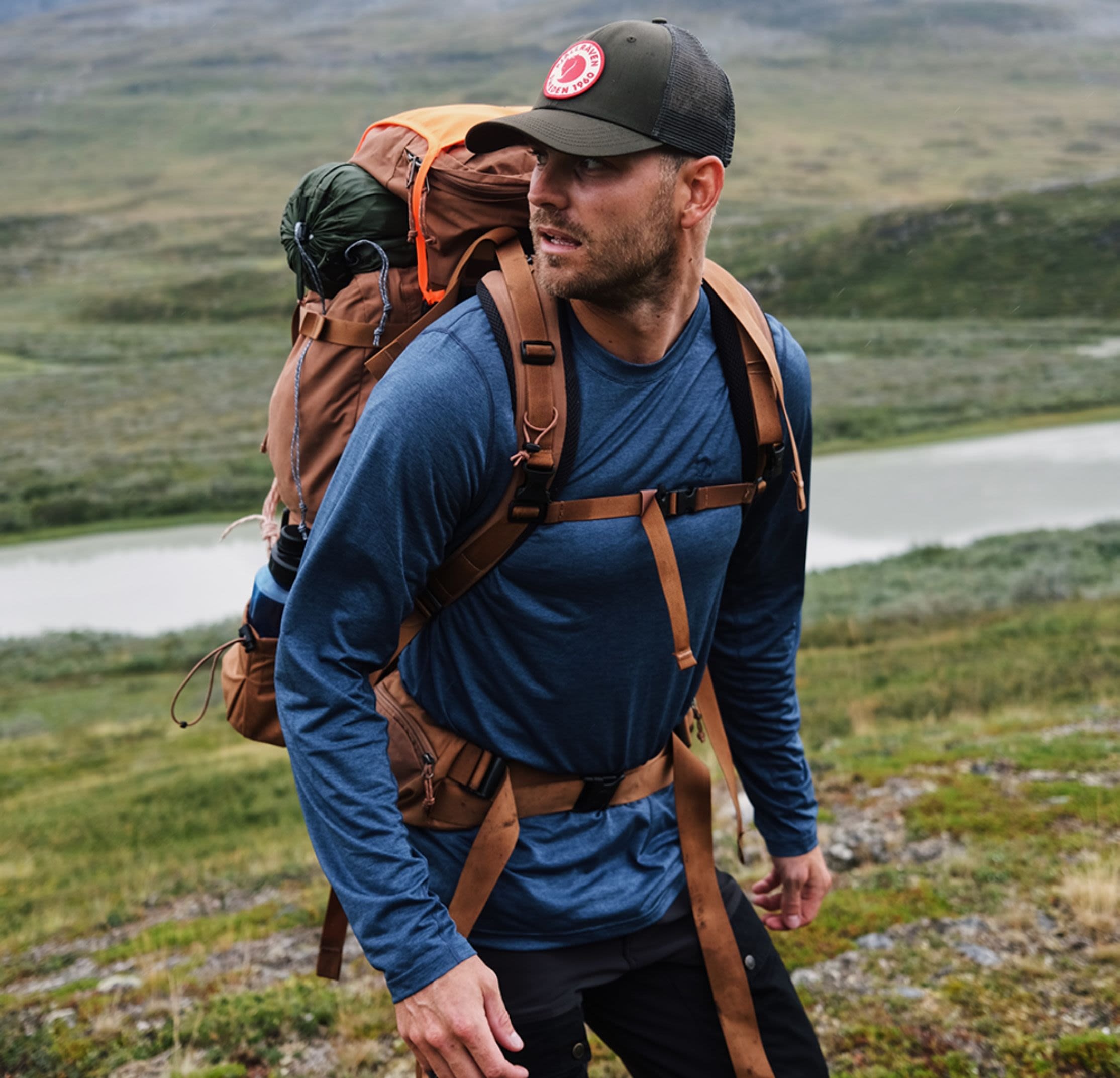Utomhusryggsäck i brunt med flera remmar, en stor grön och orange rullad sovsäck, bärs av en man i en blå långärmad skjorta.