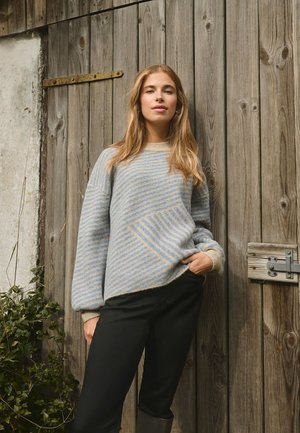 Un pull en tricot rayé bleu clair et beige avec une coupe carrée et des épaules tombantes, associé à un pantalon noir, sur un fond en bois.