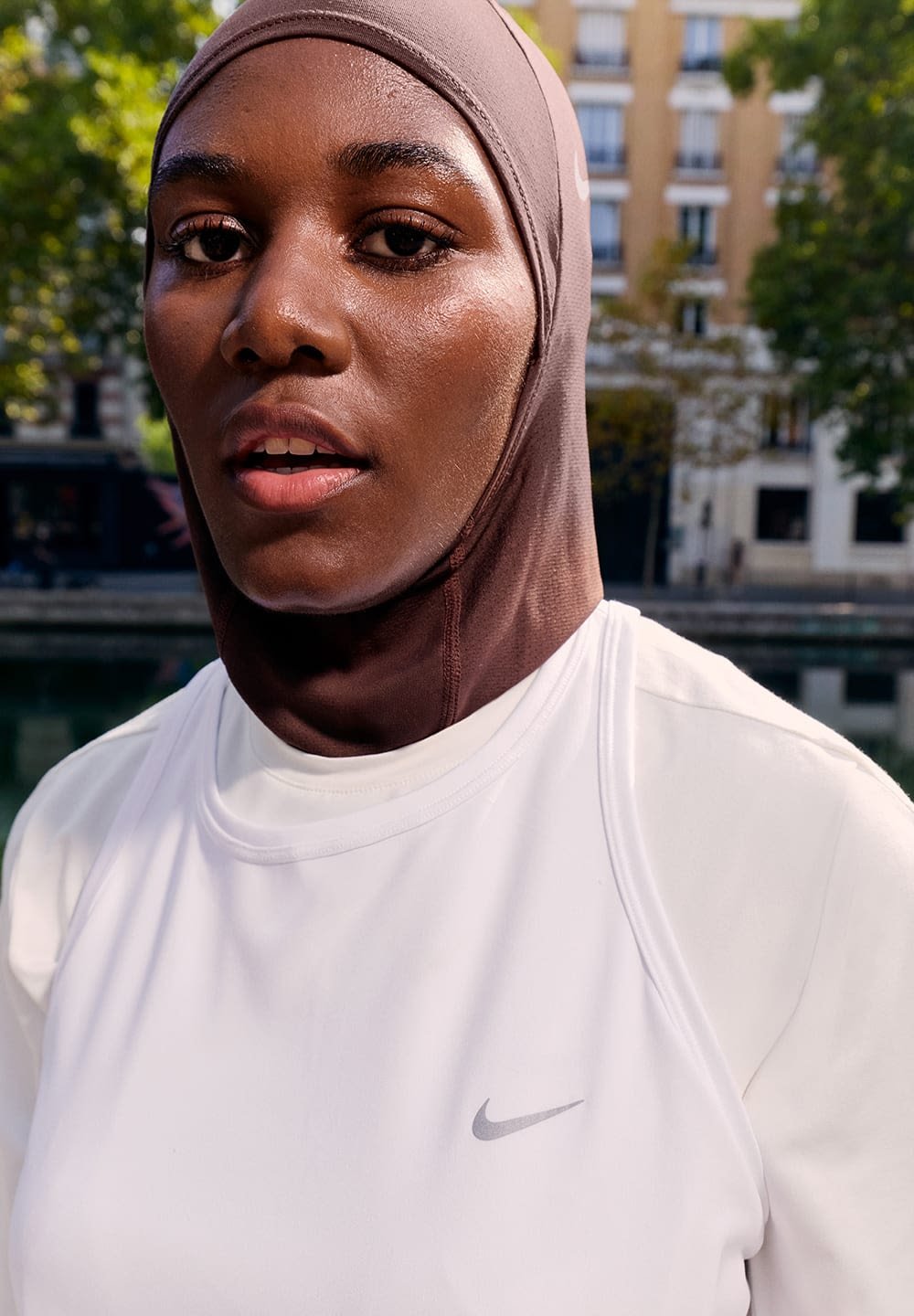 Maglietta sportiva bianca Nike con logo grigio e hijab marrone, caratterizzata da una texture liscia. Messa a fuoco sul volto, con uno sfondo sfocato.