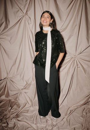 Sort palietteret top med korte ærmer, kombineret med højtaljede sorte bukser. Et hvidt tørklæde fremhæver halsudskæringen, sat mod en krøllet beige baggrund.