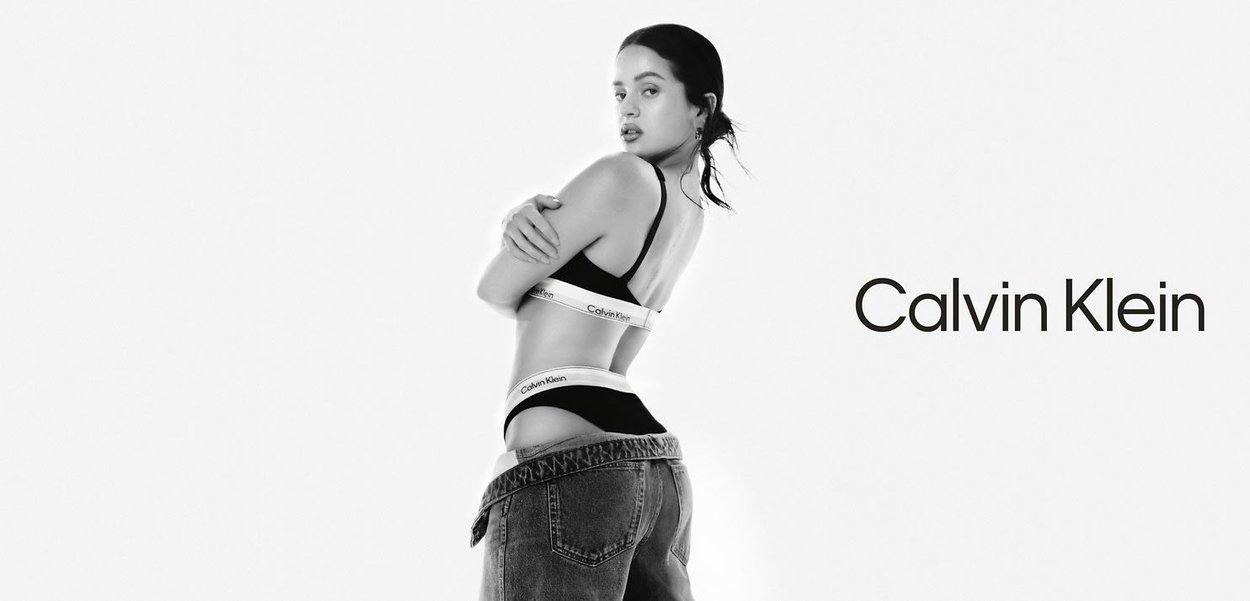 Schwarze Calvin Klein Bralette mit Logo-Band, kombiniert mit hochgeschnittenen Denim-Jeans. Das Modell posiert seitlich vor einem weißen Hintergrund.