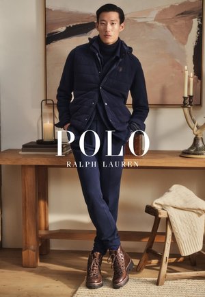 Man in marineblauwe donzen bodywarmer, coltrui en broek leunt tegen een houten tafel met een brandende kaars en een abstract schilderij in een gezellige kamer.