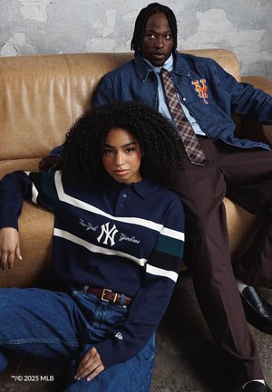Giovane donna con maglione dei New York Yankees seduta per terra appoggiata a un divano color cuoio, uomo con giacca blu dei New York Mets reclinato sul divano dietro di lei.