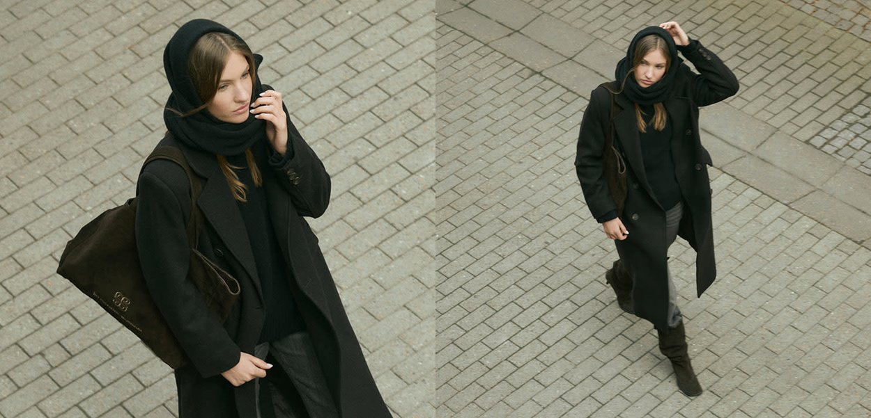 Svart kappa och halsduk, grå tröja, mörka byxor och bruna stövlar. Brun väska med logotyp. Belagd gata i bakgrunden med geometriskt mönster.