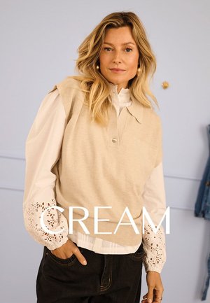 Beige gestrickte Weste mit Kragen, getragen über einer weißen Bluse mit Spitzenakzenten an den Ärmeln. Dunkle Jeans vervollständigen das Outfit.