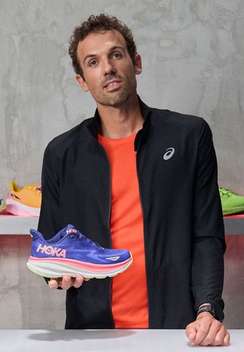 Blauer Sportschuh mit roten Akzenten und weißer Sohle, mit strukturiertem Obermaterial und prominentem HOKA-Logo. Ein Mann in schwarzer Jacke hält den Schuh.