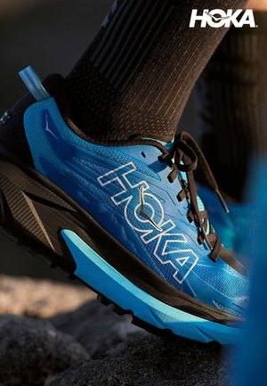 Chaussure de course Hoka bleue et noire avec des lacets noirs et de hautes chaussettes noires marchant sur un terrain rocailleux.