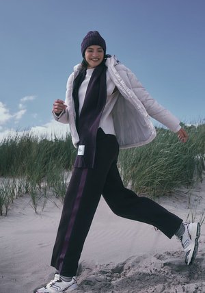 Witte pufferjack, geribbelde witte trui, zwarte broek met paarse strepen, paarse gebreide muts en bijpassende sjaal, gedragen op een zandstrand.