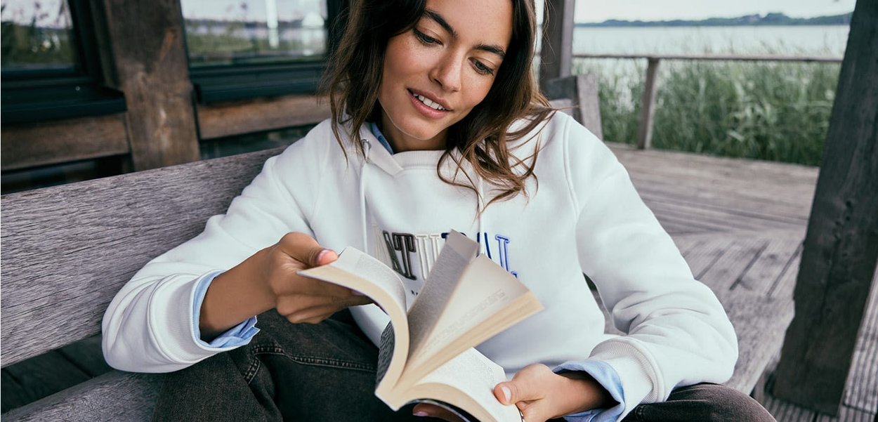 Une personne portant un hoodie blanc avec des accents bleus est assise sur un banc en bois, en train de lire un livre, entourée de hautes herbes et d'un arrière-plan serein.