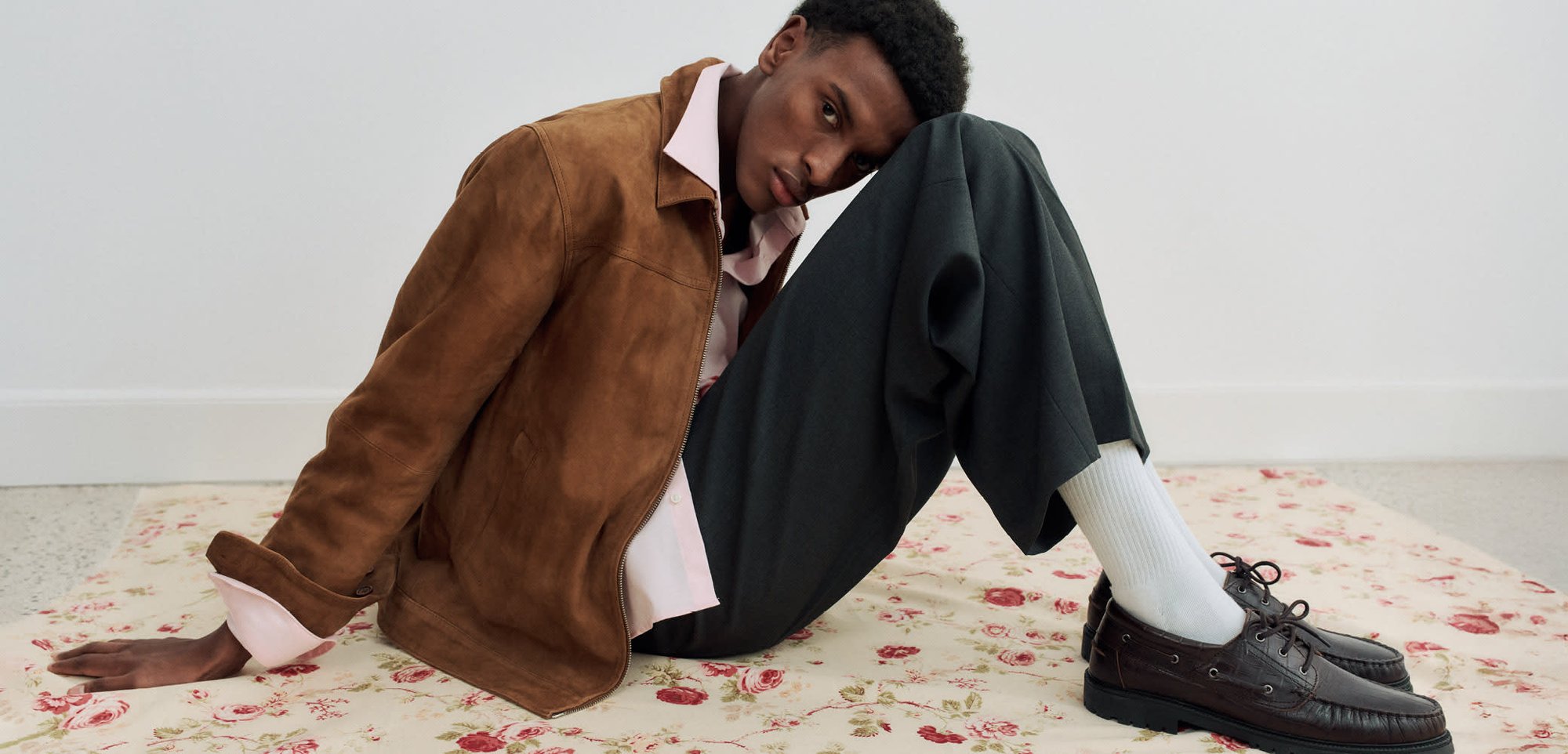 Ung man i brun mockajacka, ljusrosa skjorta, mörka byxor, vita strumpor och bruna skor som sitter på blommig matta och lutar huvudet mot knät.