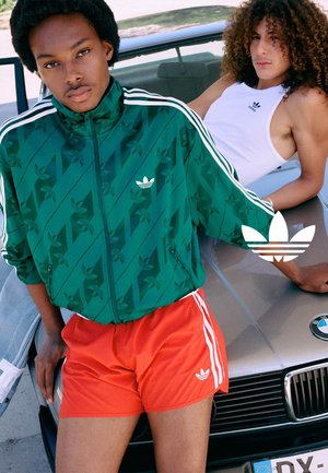 Zwei Personen lehnen an einer silbernen BMW-Motorhaube und tragen Adidas-Sportbekleidung, eine in einer grünen Jacke und roten Shorts, die andere in einem weißen Tanktop.