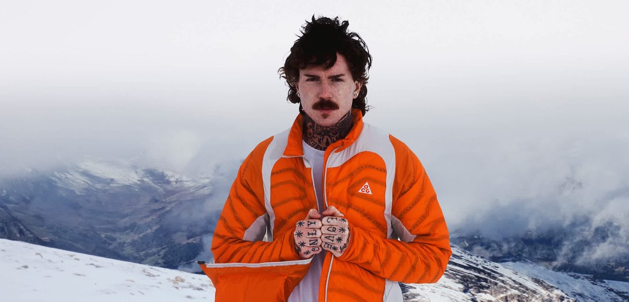 Hombre con bigote y tatuajes que lleva una chaqueta naranja y blanca, de pie en un paisaje montañoso nevado con cielo nublado.