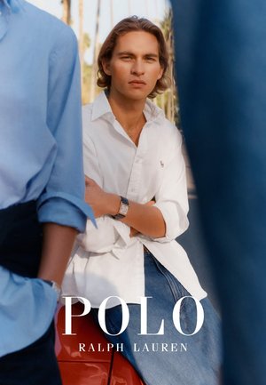 Joven vestido con camisa Polo blanca y jeans, sentado con los brazos cruzados, flanqueado por dos personas parcialmente visibles con camisas azules al aire libre, con palmeras de fondo.