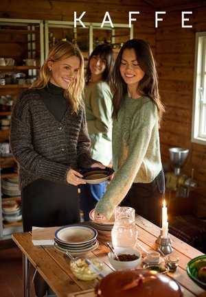 Trois femmes interagissent autour d'une table en bois avec des assiettes et de la nourriture. L'une porte un cardigan marron texturé, une autre un pull vert clair.