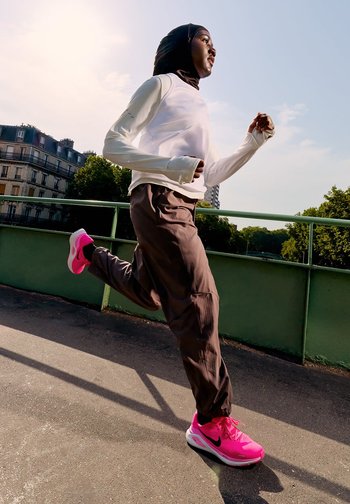 Outfit sportivo con una maglietta bianca a maniche lunghe, pantaloni marroni e scarpe da ginnastica rosa brillante con dettagli neri. Railing verde e alberi sullo sfondo.