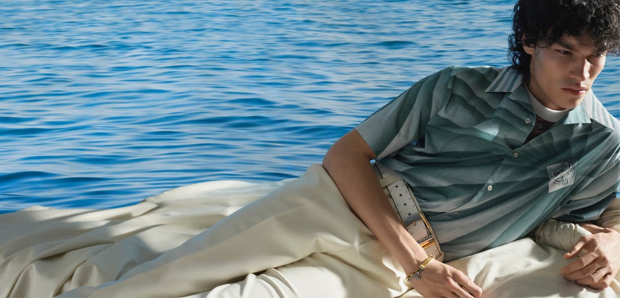 Giovane uomo con capelli ricci, sdraiato su un tessuto chiaro vicino a un mare blu calmo, indossa una camicia verde a righe e pantaloni beige con cintura.