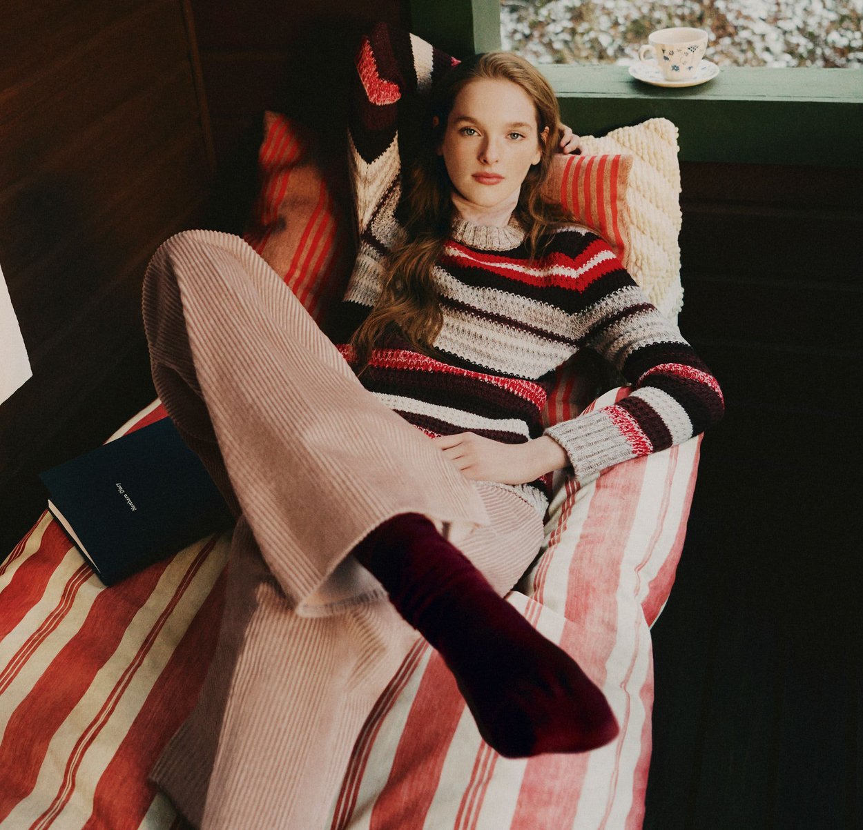 Stickad tröja med röda, grå och svarta ränder; vida manchesterbyxor i ljusrosa; bordeauxröda strumpor; randigt sängkläder; blå bok i närheten.