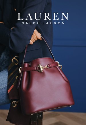 Persona sosteniendo un bolso de cuero burdeos con hebilla dorada, vistiendo un blazer azul marino y jeans azules, sobre un fondo azul oscuro.