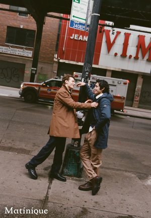 Two people interact near a street lamp. One wears a tan coat, blue pants, and black boots; the other wears a dark jacket and beige pants.