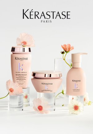 Botellas y tarro de cuidado capilar Kérastase Paris Gloss Absolu con flores rosadas claras sobre soportes acrílicos transparentes contra un fondo blanco.