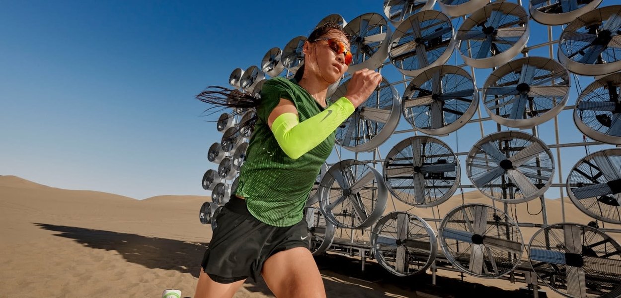 Mujer con ropa deportiva verde y mangas de brazo neón corre en el desierto cerca de una gran serie de ventiladores industriales en una estructura metálica.