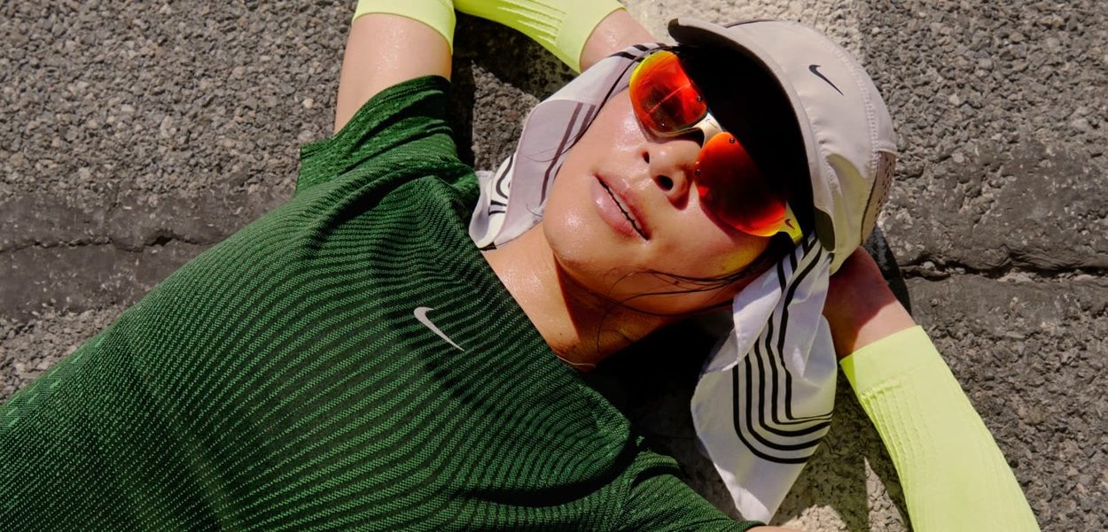 Persona con abbigliamento sportivo verde e cappellino bianco sdraiata a riposare su un marciapiede ruvido, indossa occhiali da sole riflettenti rossi e manicotti giallo neon.