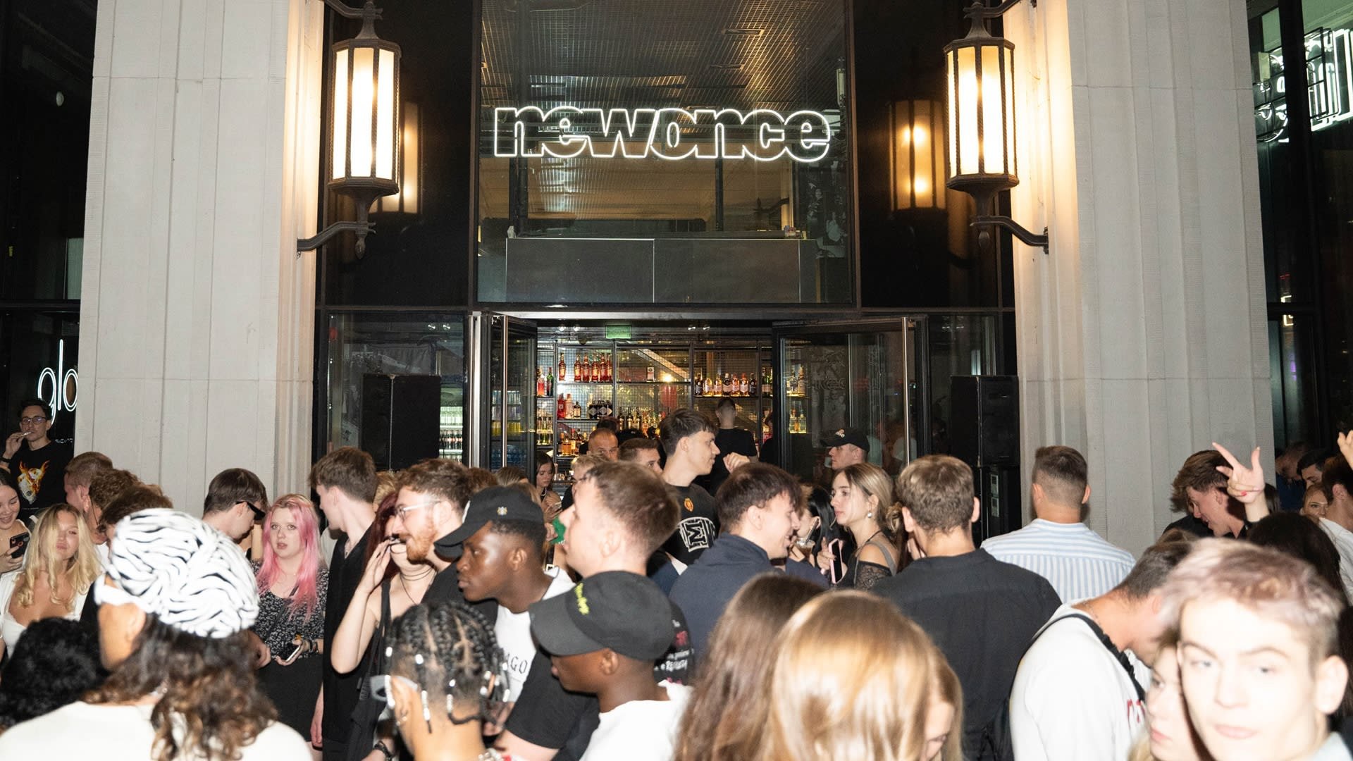 Een menigte verzameld buiten een bar met een glazen façade en verlichte lichtreclame. Opvallende ronde verlichting en donkere pilaren omlijsten de ingang.