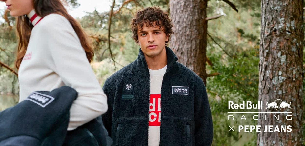 Giacca in pile nera con tasche frontali e loghi, con un colletto alto. Lo sfondo include alberi e un lago, suggerendo un ambiente all'aperto.