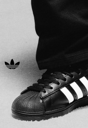 Scarpa da ginnastica Adidas nera con strisce bianche e punta texturizzata, indossata con pantaloni neri, logo trefoil Adidas vicino su sfondo grigio.