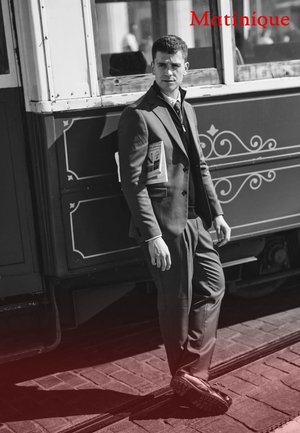 Grauer Anzug mit strukturiertem Design, passendem Hemd und Krawatte. Hält eine Zeitung und steht neben einer nostalgischen Straßenbahn auf einer Kopfsteinpflasterstraße.