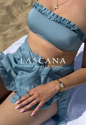 Frau trägt hellblaue, mit Rüschen versehene Bikini und passenden Sarong auf einem Strandtuch mit einer goldenen Uhr und Halskette.