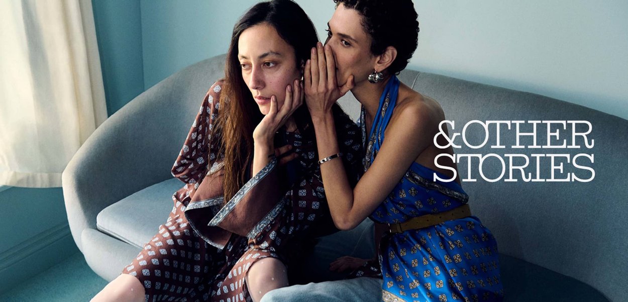 To kvinder sidder på en grå sofa; den ene hvisker i den andens øre, begge iført mønstrede kjoler i et blødt oplyst rum.