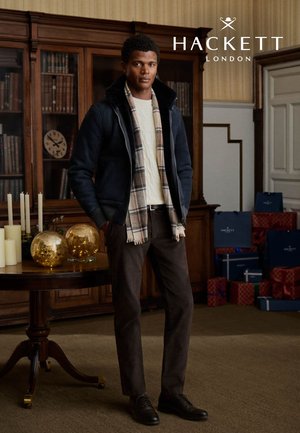 Marineblauw jack met een hoge kraag, crème trui, geruite sjaal, bruine broek en donkere schoenen. Gouden glazen bollen en witte kaarsen op een tafel.