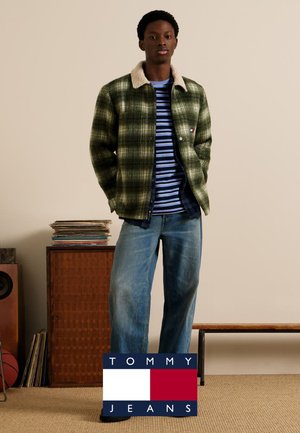 Giacca a quadri verde con colletto bianco, indossata sopra una camicia a righe blu e bianche, abbinata a jeans blu dal taglio morbido.