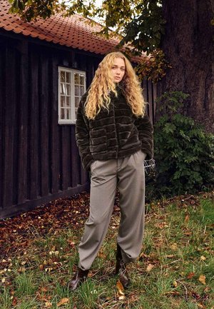 Faux fur olive green jacket with textured, quilted pattern paired with light gray, loose-fitting trousers and brown ankle boots in an outdoor setting.