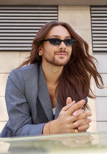 Sunglasses with a narrow, cat-eye shape and black frame. Model wears a gray blazer, appears with long hair and layered accessories.