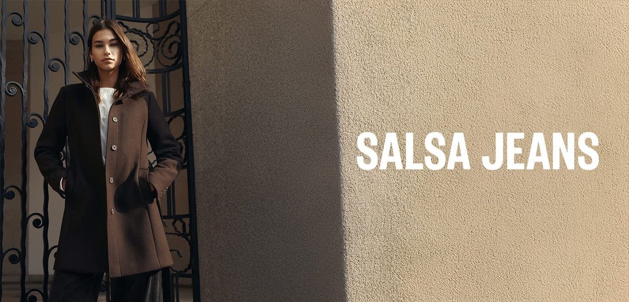 Brauner Mantel mit hohem Kragen, Knöpfen und seitlichen Taschen, getragen über einem weißen Oberteil, vor einem strukturierten beigen Hintergrund.