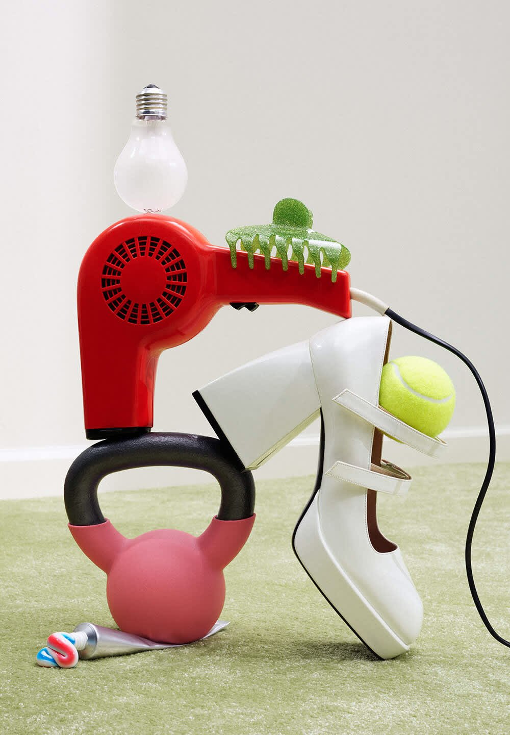 Ein roter Haartrockner mit einer Glühbirne oben drauf, neben einem weißen Schuh, einem grünen Tennisball, einem pinken Kettlebell und bunten Röhren auf einer grünen Fläche.