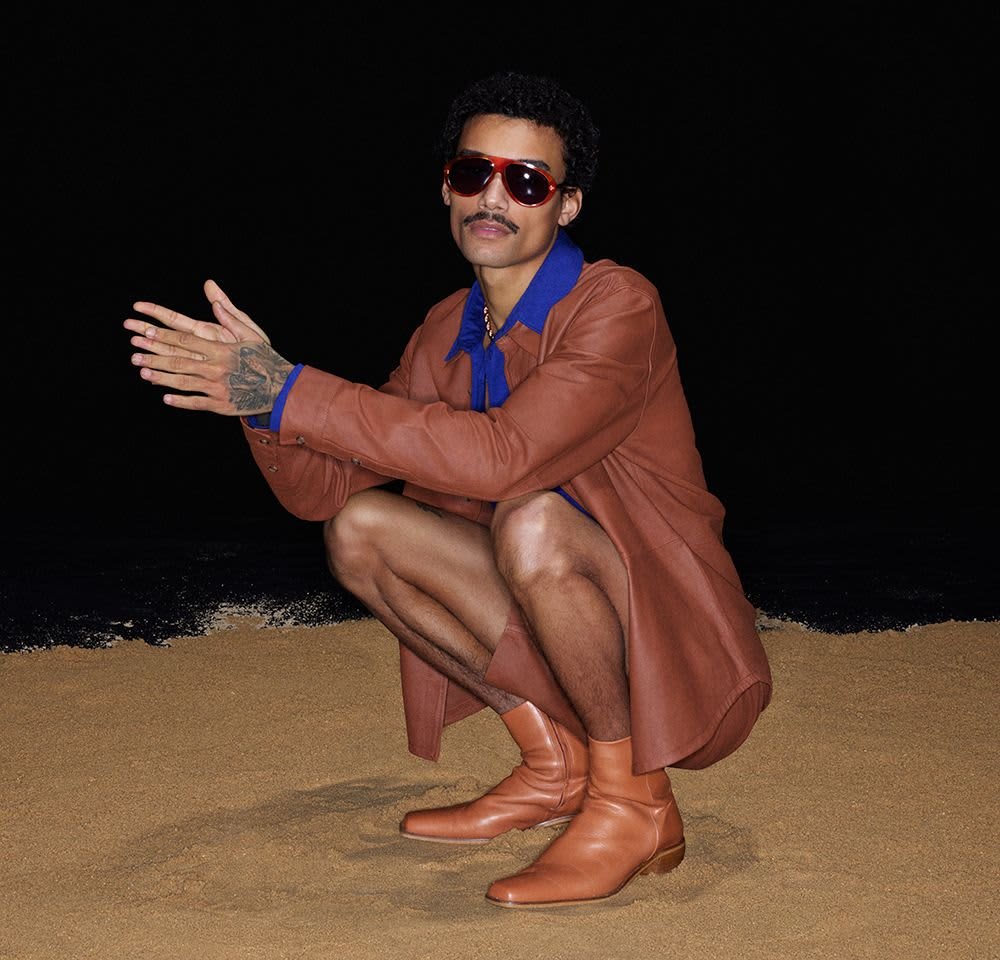 Brown leather overshirt, blue collar, red sunglasses, and brown ankle boots. Model crouching on sand, showcasing smooth textures and vibrant colours.