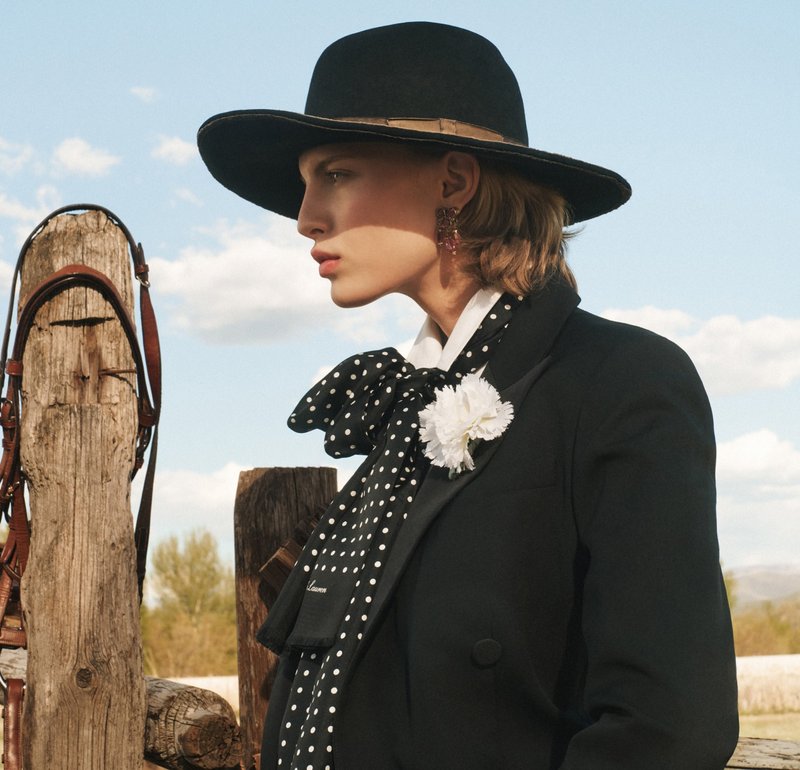 Chapeau en feutre noir, écharpe à pois, blazer noir formel et accent floral blanc. Pieux en bois en arrière-plan avec des sangles en cuir visibles.