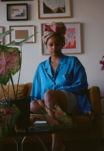 Blauwe satijnen pyjamashirt met een textuurpatroon, gedragen over grijze shorts. Heeft een roze turban en een groene plant in een zwarte pot in de buurt.